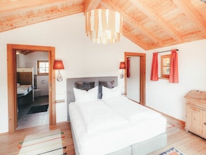 A bedroom with a wooden ceiling, a bed with white bedding, a wooden cabinet, and a view into the bathroom. - Luna Blanca Chalet (Schwangau)