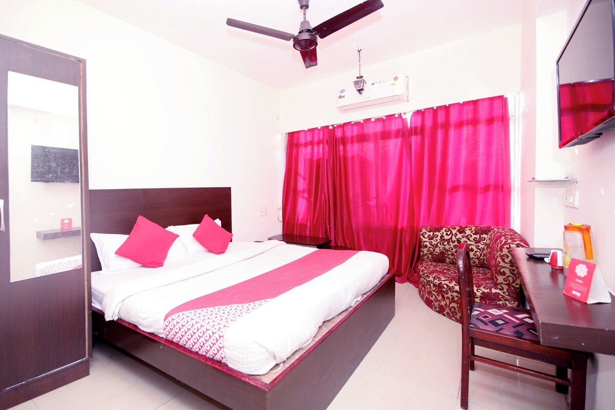 Une chambre d’hôtel avec un lit, une chaise, un bureau, un ventilateur de plafond et une fenêtre avec des rideaux rouges.