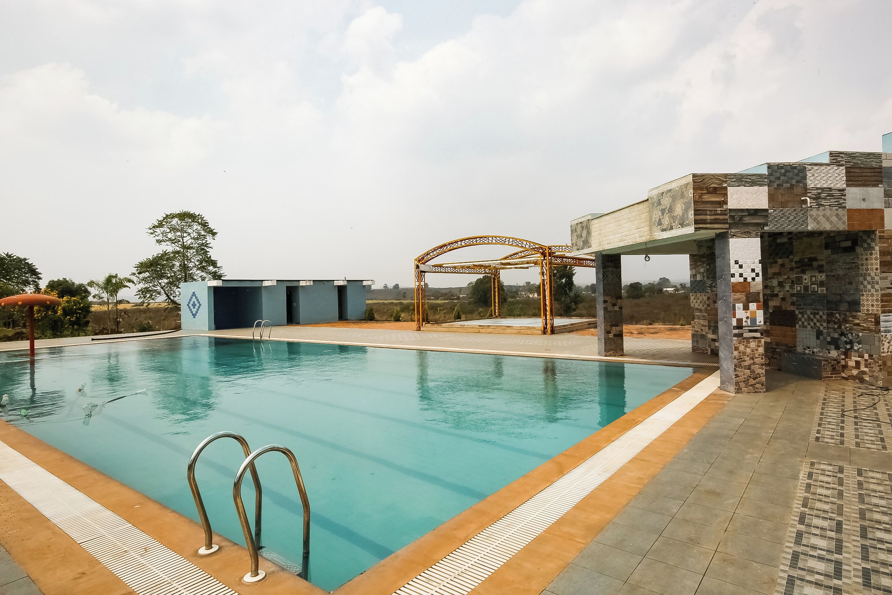 A swimming pool with a metal ladder, a covered seating area, and a building with mosaic tilework.