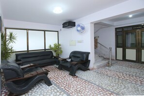 A lobby with a reception desk, a sofa, a chair, a potted plant, a wall-mounted fan, and a staircase leading to a restaurant. - Hotel O Sukooon City View (Jabalpur)
