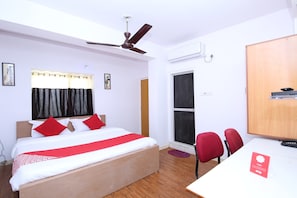 Une chambre d’hôtel comprenant un lit, un bureau, une chaise, une armoire, un ventilateur de plafond, un appareil de climatisation et une fenêtre avec des rideaux.