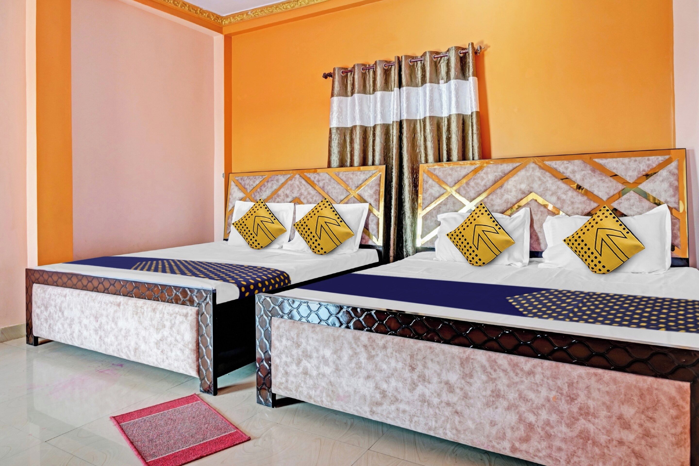 A hotel room with two beds, each with a patterned headboard, a window with curtains, and a red doormat.