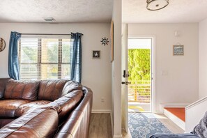 Ett vardagsrum med en brun lädersoffa, ett fönster med blå gardiner, en stjärndekoration och en dörr som leder ut till en balkong med ett trästaket.
