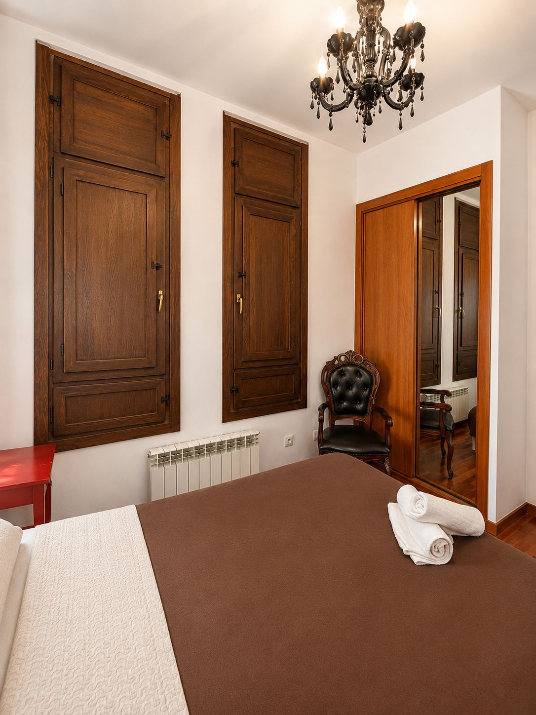 Un dormitorio con una cama grande, un armario de madera, un radiador, una silla y una araña.