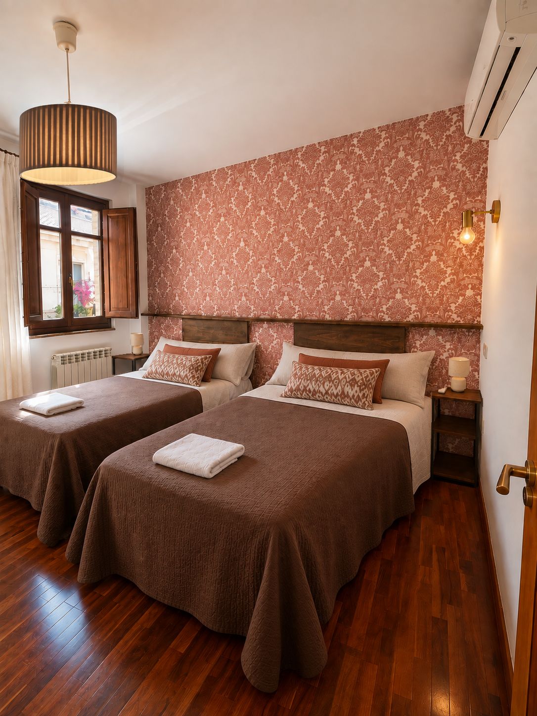 Habitación de hotel con dos camas, una ventana con cortinas, un radiador, una pared con papel tapiz estampado y una lámpara colgante.