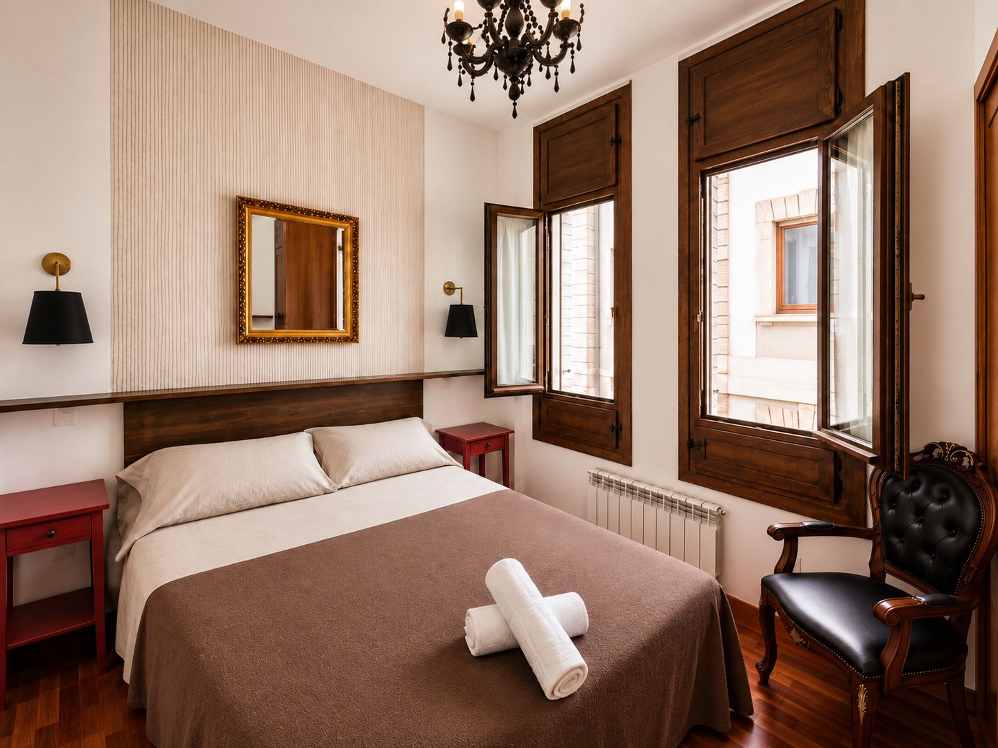 A hotel room with a large bed, two bedside tables, a mirror, a chandelier, and a window with shutters.
