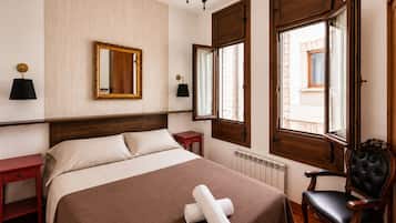 A hotel room with a large bed, two bedside tables, a mirror, a chandelier, and a window with shutters.