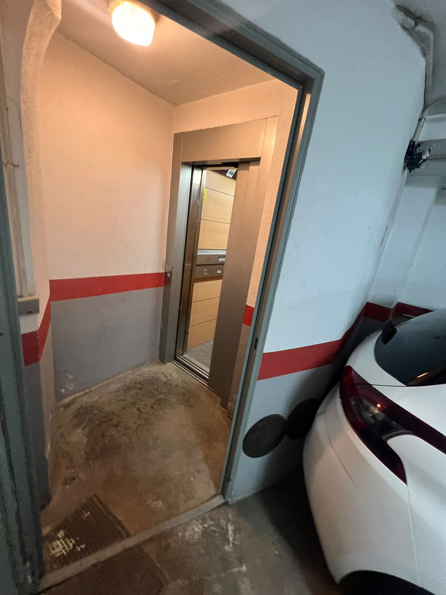 En smal hall med en vit bil parkerad på höger sida, en dörr som leder in till ett rum med en röd rand, och en lampa i taket.