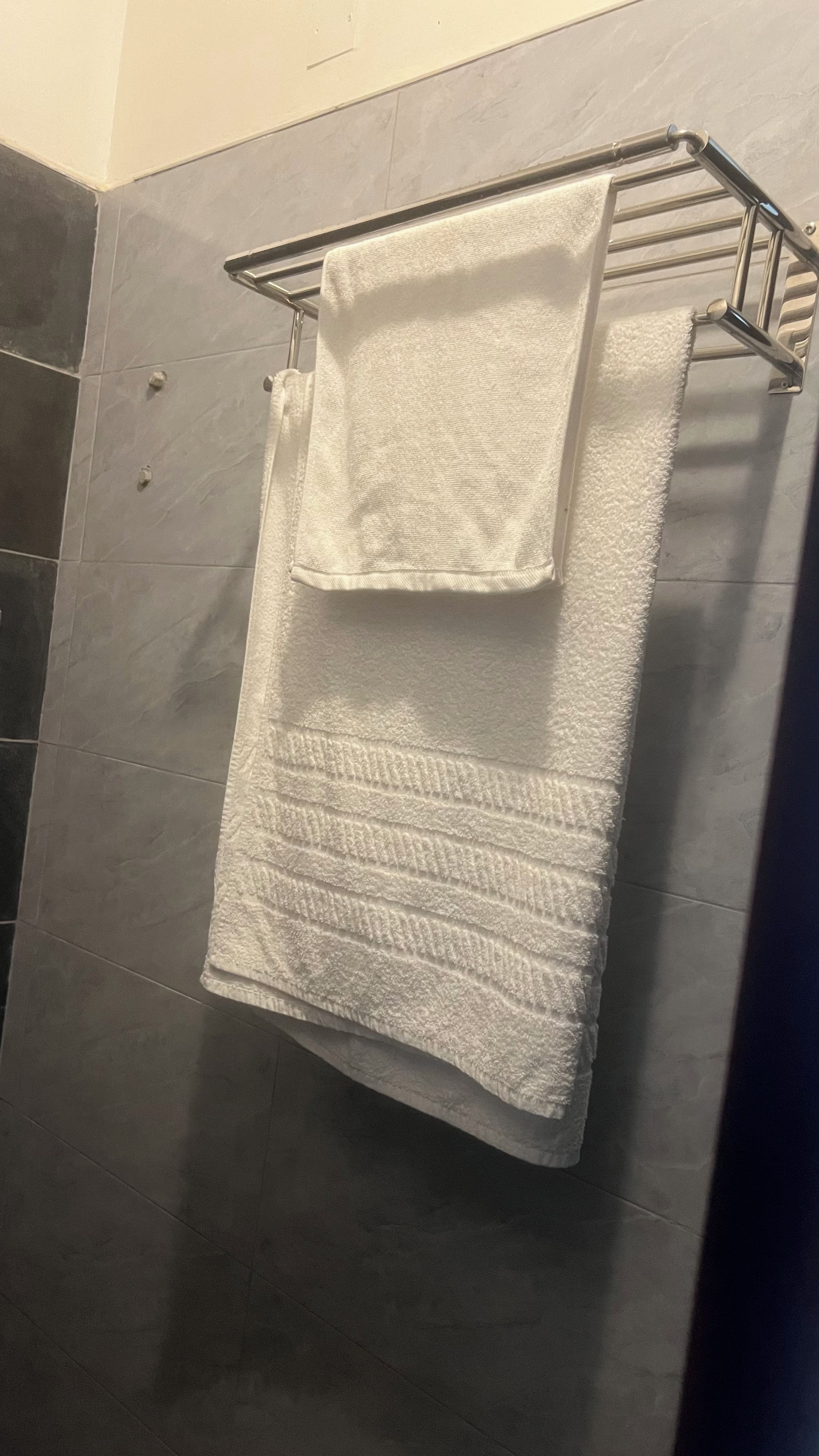 Una toalla blanca colgada en un tendedero metálico en un baño con paredes de azulejos oscuros.