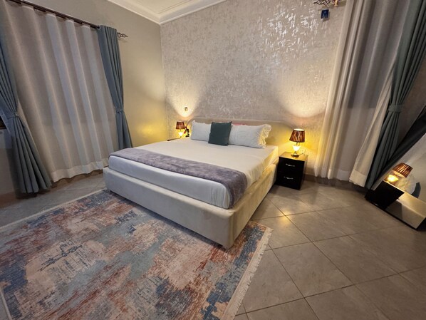 Un dormitorio con una cama grande, lámparas de noche, una alfombra estampada y cortinas.