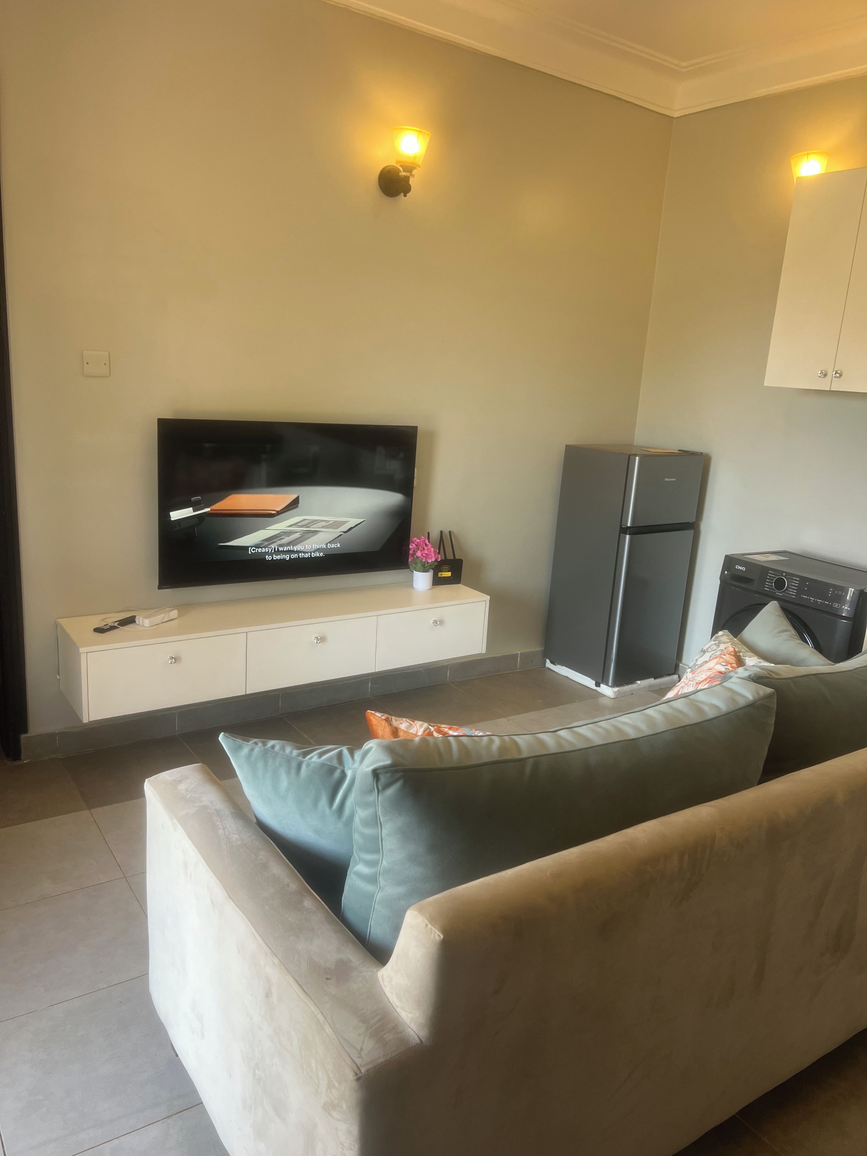 Una sala de estar con televisor de pantalla plana, refrigerador y sofá con cojines.