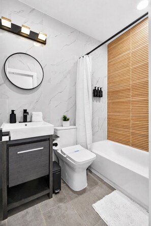 Ein modernes Badezimmer mit Marmorwand, Jalousie aus Holz, einer weißen Badewanne und einem runden Spiegel.