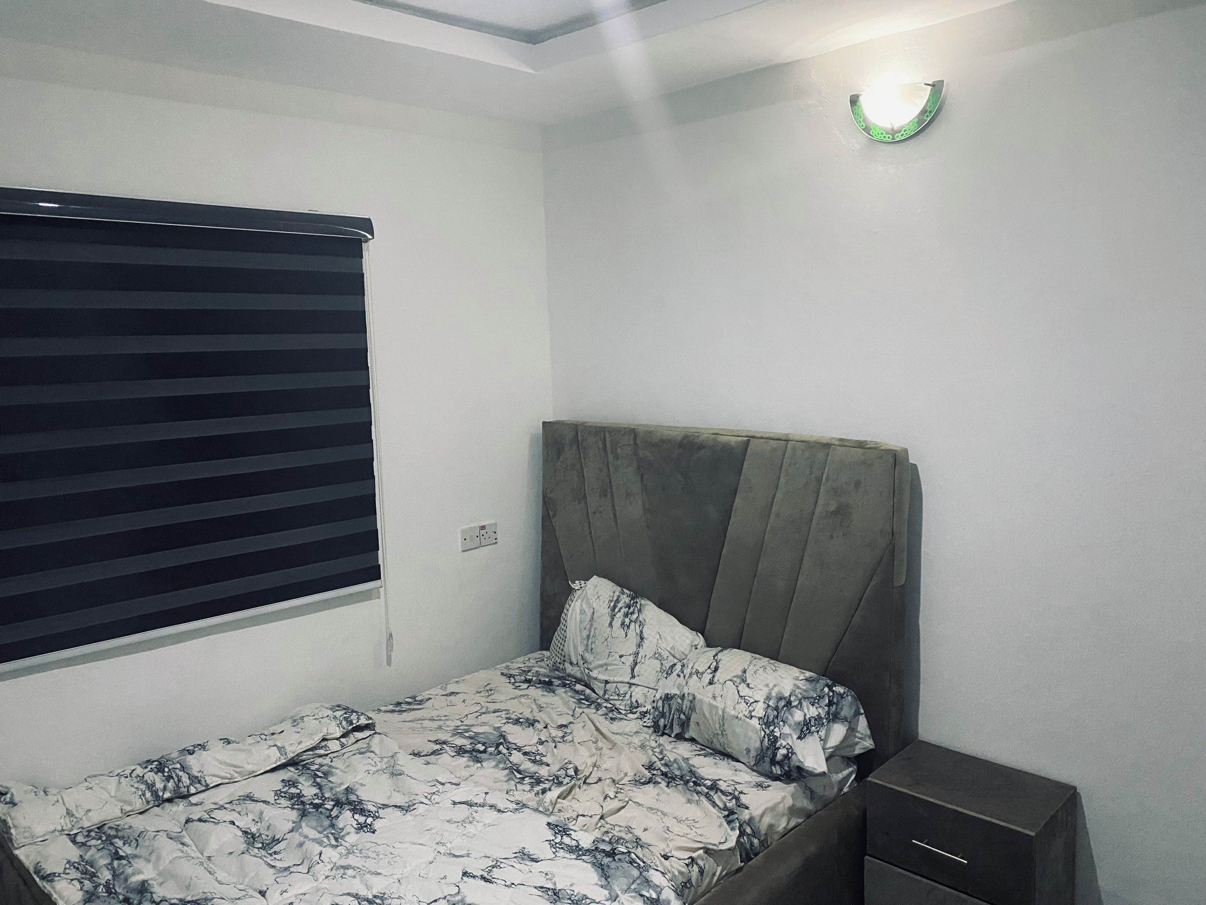 Une chambre à coucher comprenant un lit, une table de chevet, une prise électrique et une fenêtre avec des stores.
