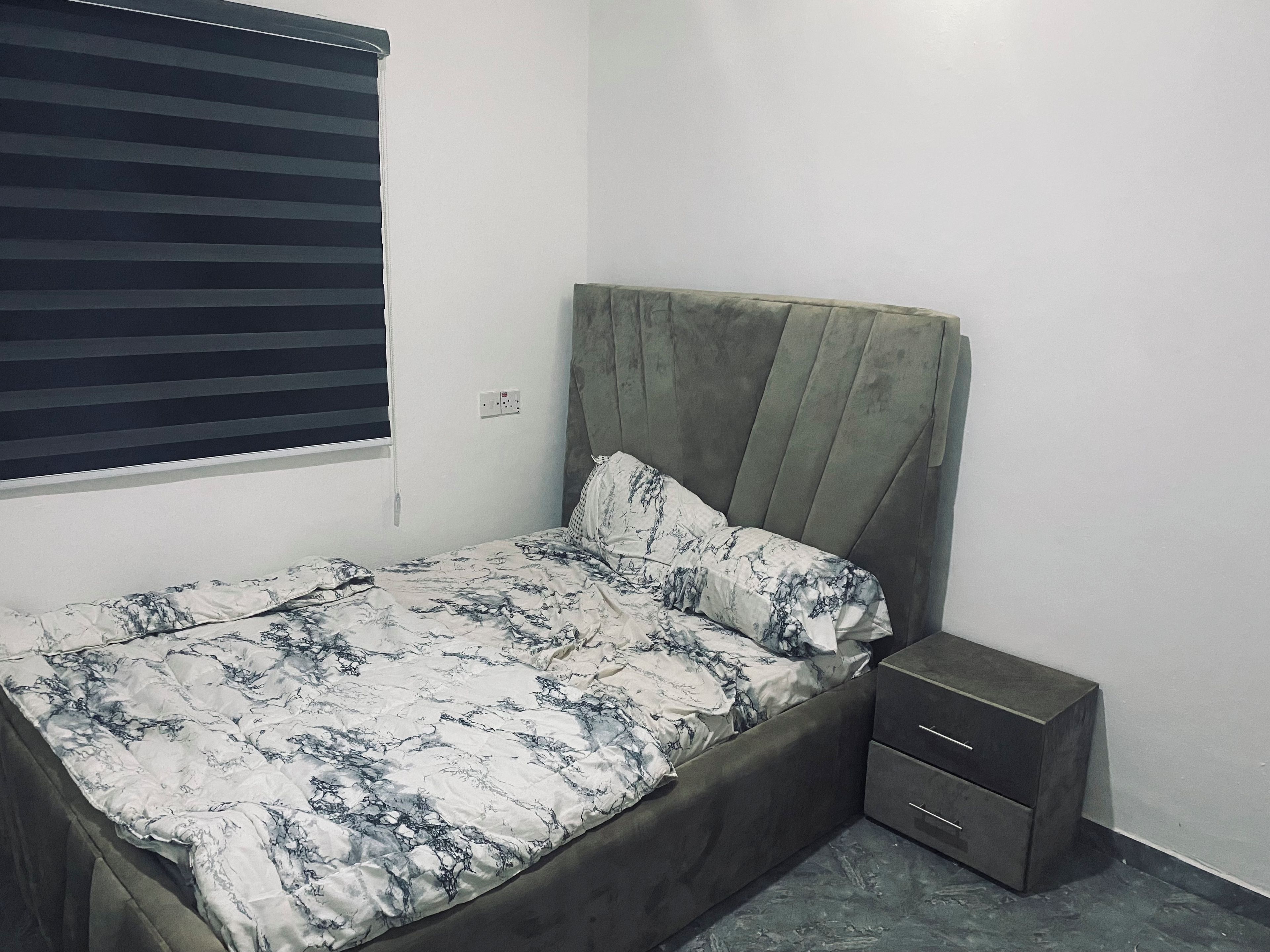 Un lit simple avec une tête de lit, une commode de chevet et une fenêtre avec des stores.