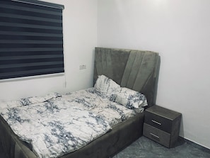 Una cama individual con cabecera, un cajón junto a la cama y una ventana con persianas.