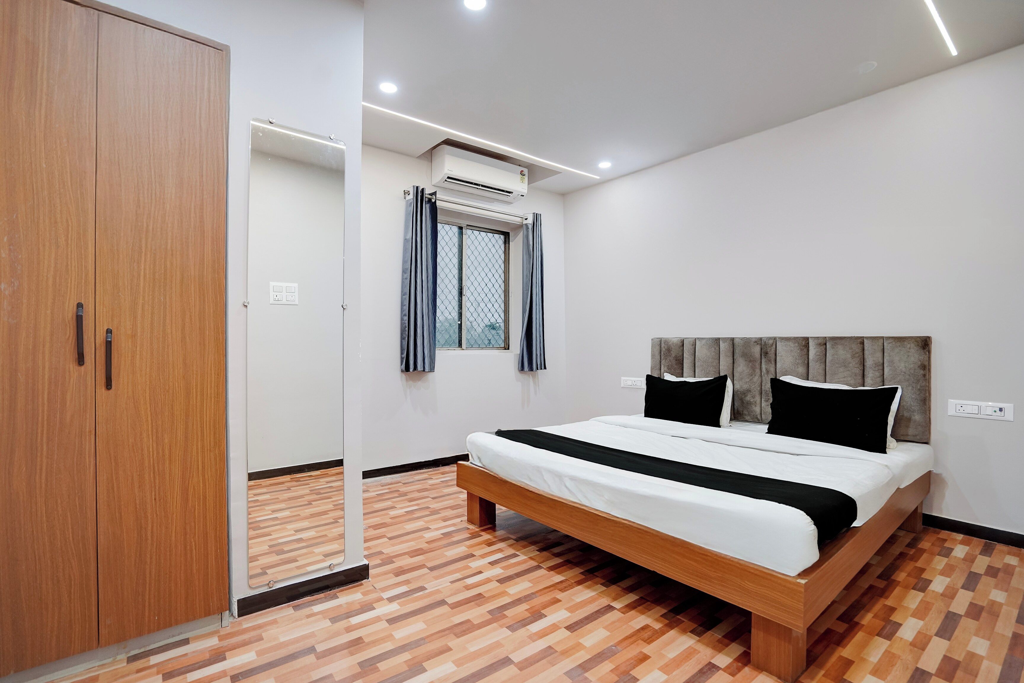 A bedroom with a wooden wardrobe, a bed with black and white bedding, a window with curtains, and an air conditioning unit.