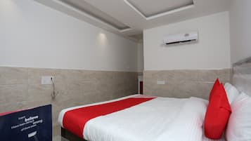 A hotel room with a bed, a red and white bedspread, a red pillow, a wall-mounted air conditioner, and a poster on the wall.