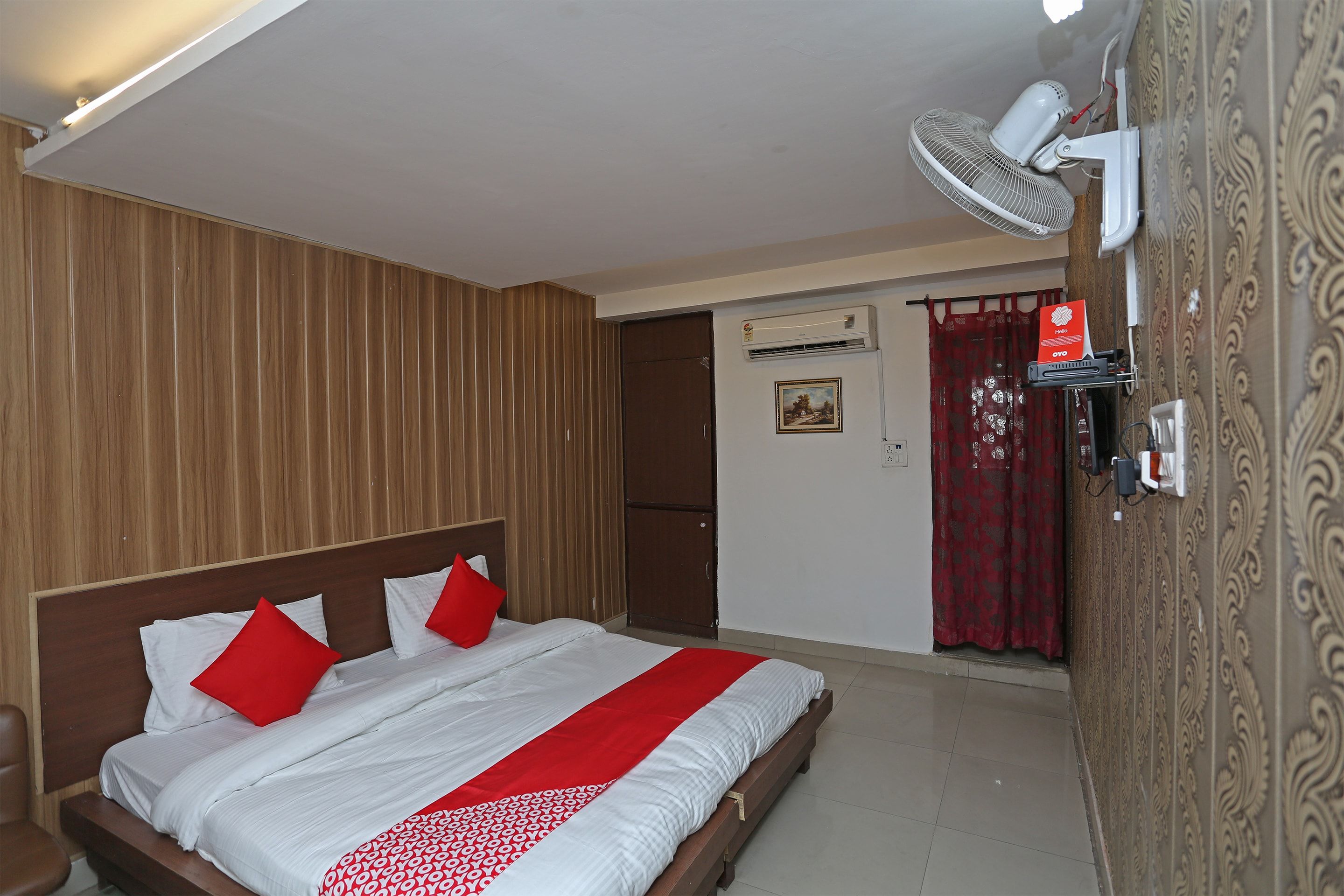 Habitación de hotel con cabecera de madera, cama con ropa blanca y detalles rojos, ventilador de pie, unidad de aire acondicionado y pared estampada.