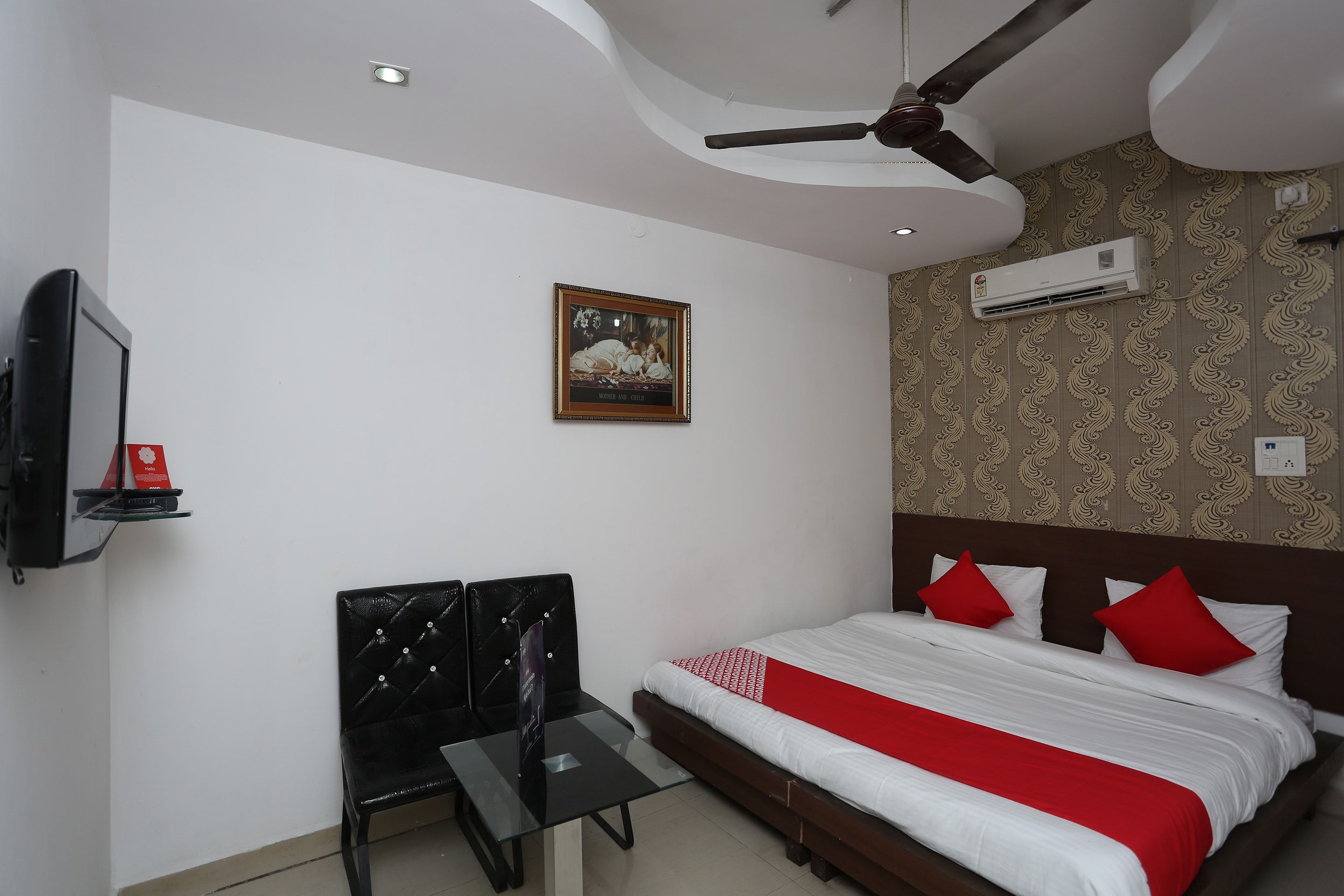 A hotel room with a bed, a desk, a chair, a ceiling fan, an air conditioning unit, and a framed picture on the wall.