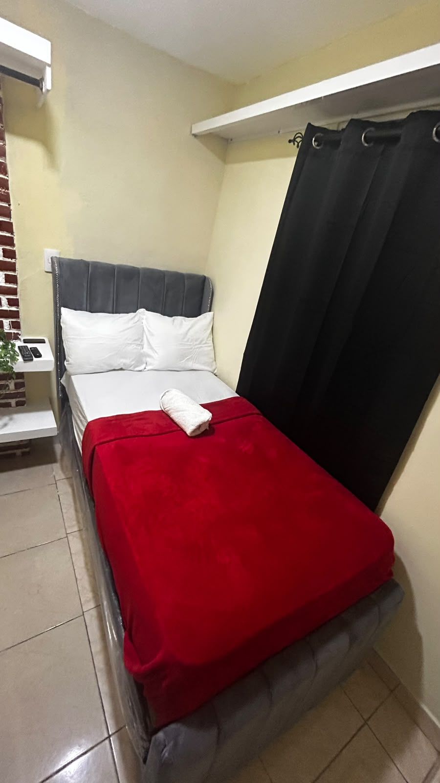 Un lit simple avec une couverture rouge, une tête de lit grise et un rideau noir.