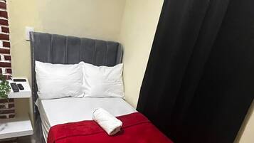 Un lit simple avec une couverture rouge, une tête de lit grise et un rideau noir.