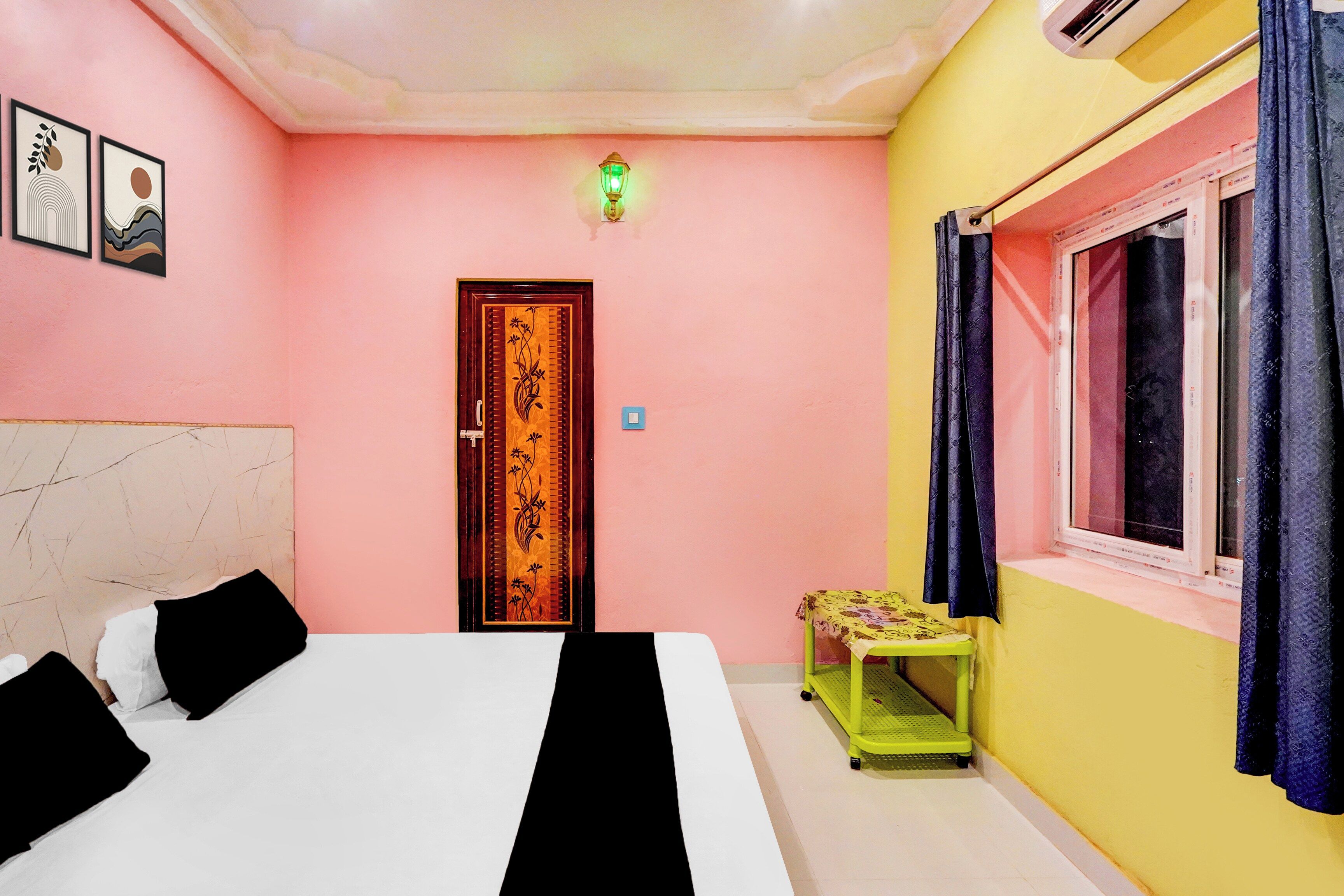 A bedroom with a bed, a door, a small table, a window with curtains, and a wall-mounted light.