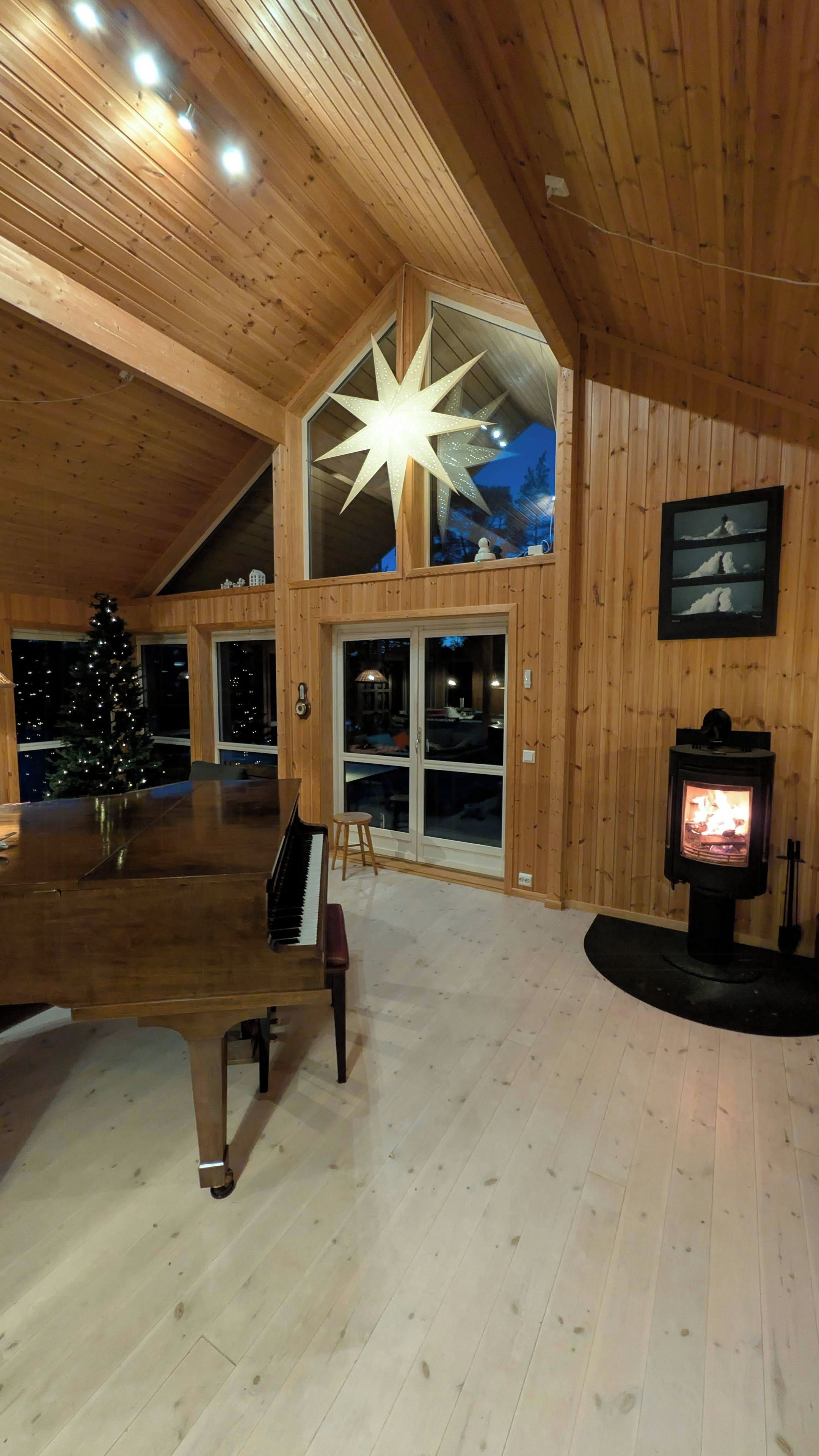 Une pièce spacieuse avec un plafond en bois, un grand luminaire en forme d’étoile, un piano, un sapin de Noël et un poêle à bois.
