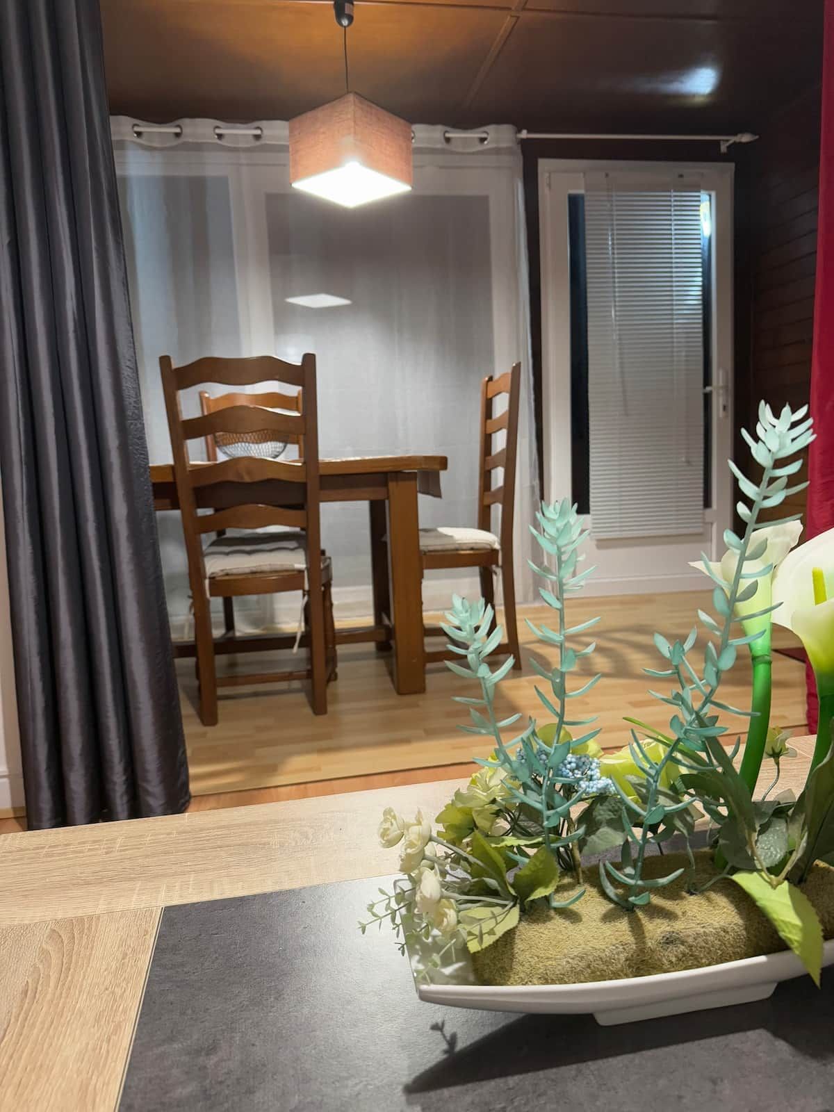 Een eethoek met een houten tafel en stoelen, een hanglamp en een potplant met kunstbloemen.