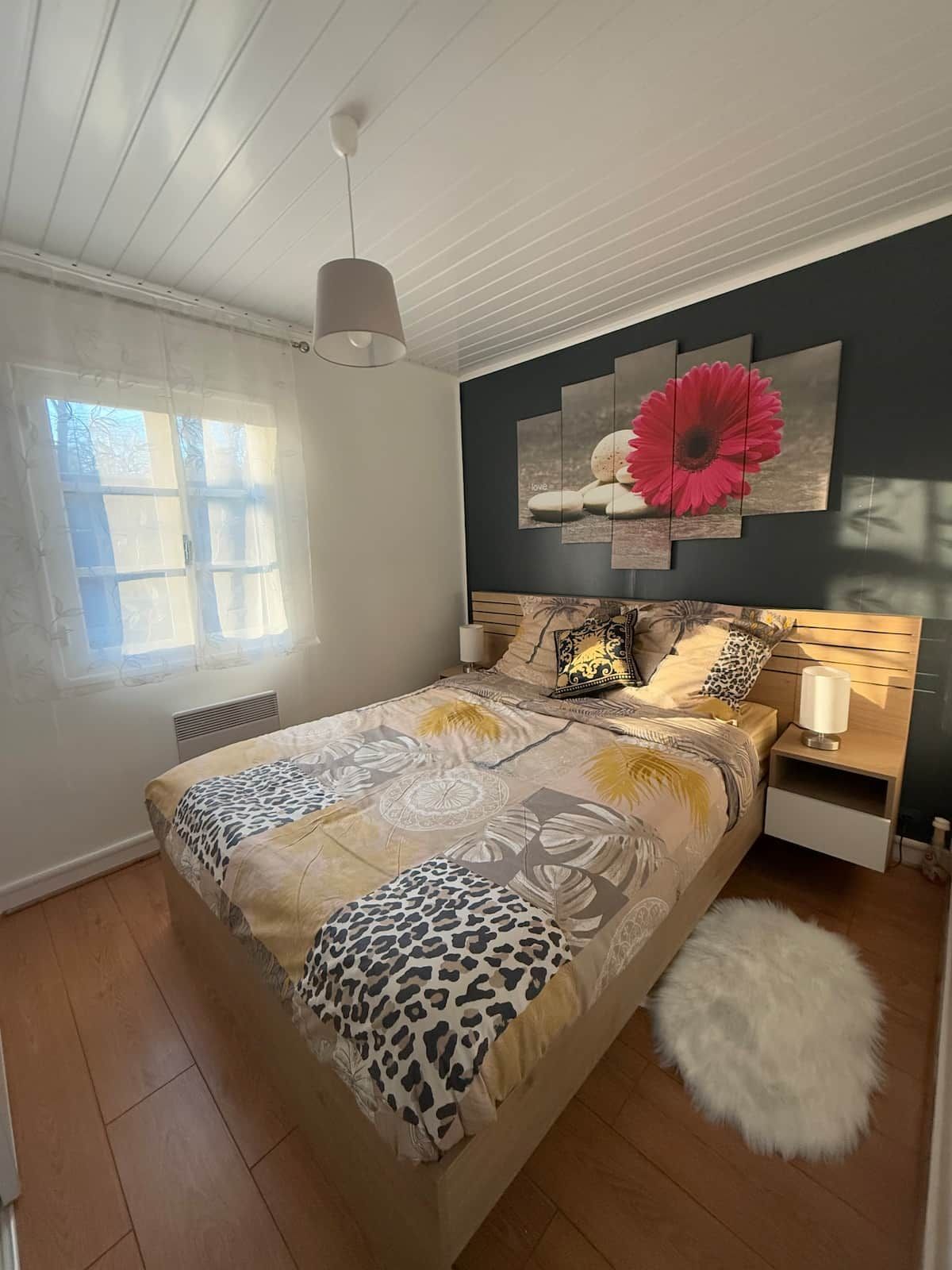 Een slaapkamer met een bed, nachtkastje, kunstwerk aan de muur en een raam met dunne gordijnen.