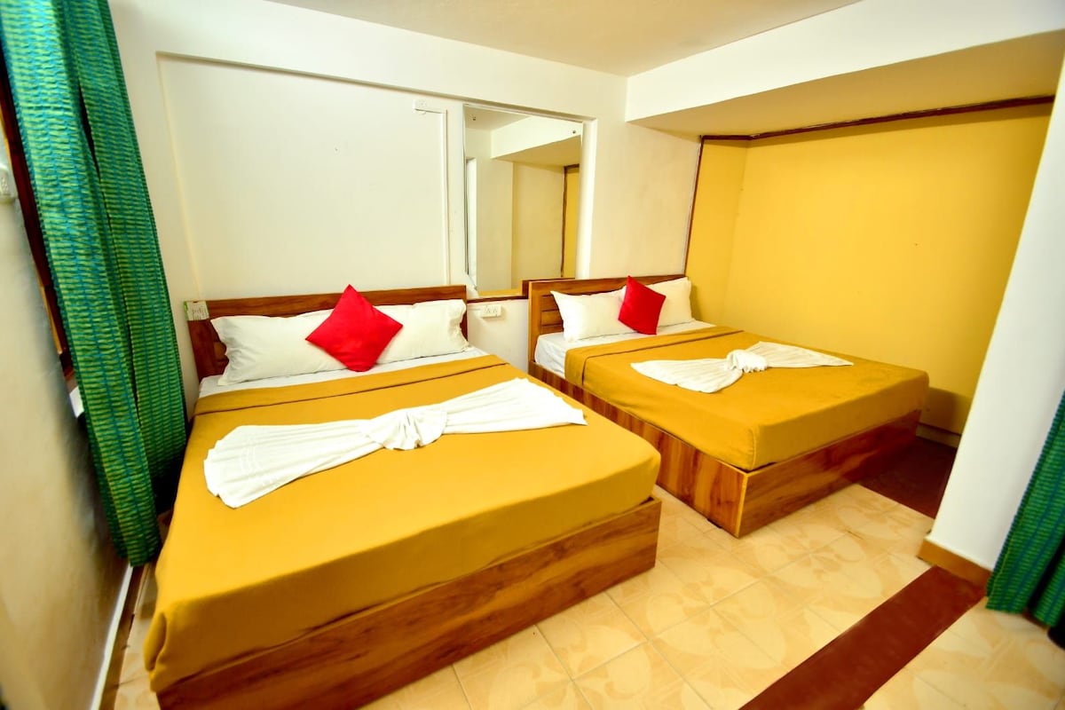 Een hotelkamer met twee bedden, elk met een houten hoofdeinde, witte beddengoed en rode kussens. Er hangt een spiegel aan de muur en een groen gordijn is gedeeltelijk zichtbaar.