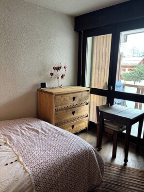 A bedroom with a bed, a dresser with heart-shaped cutouts, a small table, and a window with a view of buildings. - Appartement cosy – aux 2 Alpes (Les Deux Alpes)