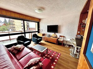 Een woonkamer met een bank, stoelen, een televisie en een eethoek met stoelen en een tafel.