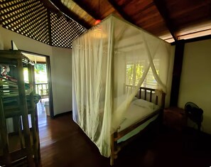 A room with a wooden bed, a wooden chair, a wooden table, and a window with a view of greenery. - Cabo Matapalo Rainforest Beach House (Puntarenas Province)