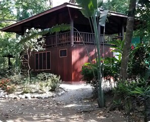 Ein Holzhaus mit einem Balkon, umgeben von dichter Vegetation und einem Kiesweg.