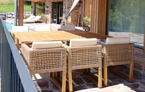 An outdoor dining area with a wooden table and wicker chairs, a stone floor, and a view of a pool and greenery. - New Listing (Lieschen)