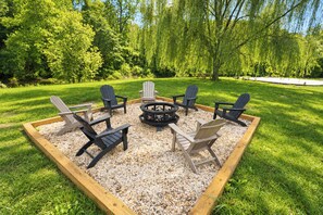 A circular seating arrangement with a central fire pit, surrounded by wooden chairs and a wooden table, set on a gravel area within a grassy landscape.