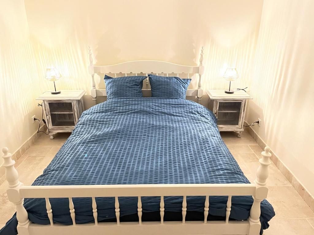 Een bed met een wit frame en blauw beddengoed, omringd door twee nachtkastjes met lampen, in een kamer met effen muren en een betegelde vloer.