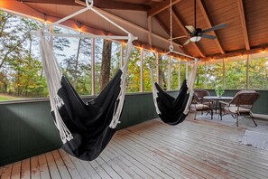 A wooden deck with two hammocks suspended from the ceiling, a table with chairs, and a view of trees through large windows. - Bluebell Cottage-Forest Views & Fireside Evenings (Luray)