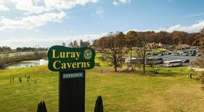 A sign for Luray Caverns with an entrance, a parking lot, and a view of a lake and buildings in the background. - Bluebell Cottage-Forest Views & Fireside Evenings (Luray)