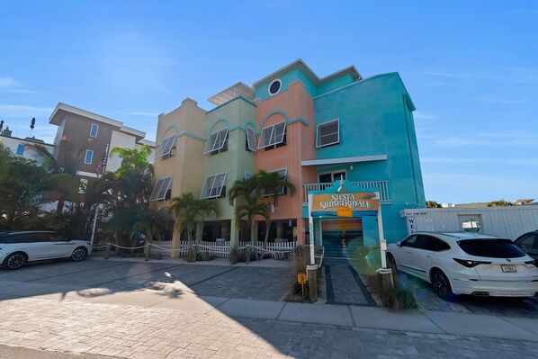 A multi-story hotel with a colorful facade, a gated entrance, and a parking area with cars. - Siesta Key | Pool, Spa & Flexible 1-2 Bedroom Lock-Off (Siesta Key)