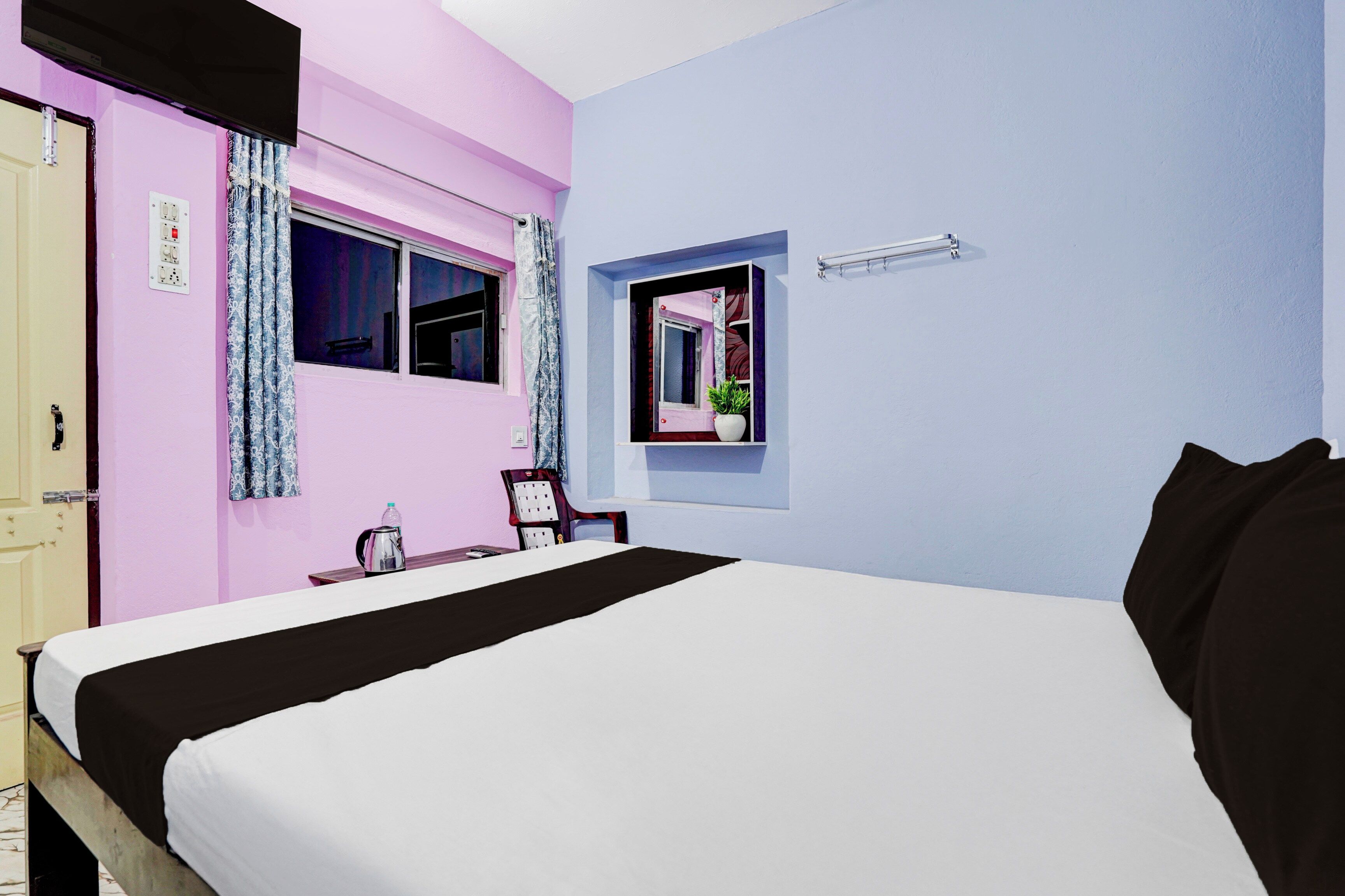 A bedroom with a bed, a window with curtains, a bedside table with a kettle, and a small potted plant.