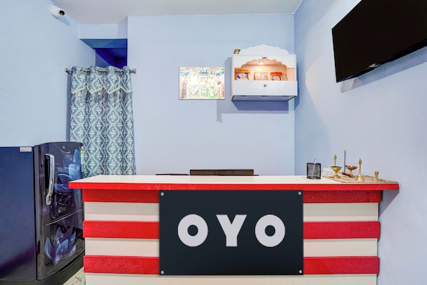 A reception desk with a sign reading 'OYO' in a room with light blue walls, a mounted television, and a small shrine with religious items.