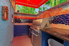 Eine kompakte Küche mit integriertem Grill, einem kleinen Spülbecken und einem Fenster mit Blick auf einen tropischen Garten.