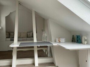 Ein weißer Schreibtisch mit Lampe und Buchstabenwürfeln in einem Raum mit geneigten Decken und einer eingebauten Sitzbank mit Kissen.