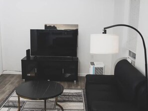 A living room with a black sofa, a black coffee table, a flat-screen TV on a black stand, a white floor lamp, and a patterned rug. - Stylish 2 bedroom apartment located in the heart of Newark; Close to NYC. (Newark)
