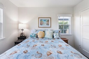 A bedroom with a bed featuring a seashell pattern, a nightstand with a lamp, a framed painting of a beach scene, and a window with a view of greenery. - Bright Coastal 2BR Home • Huge Yard • Walkable • 10 Mins To The Beach (Los Angeles)