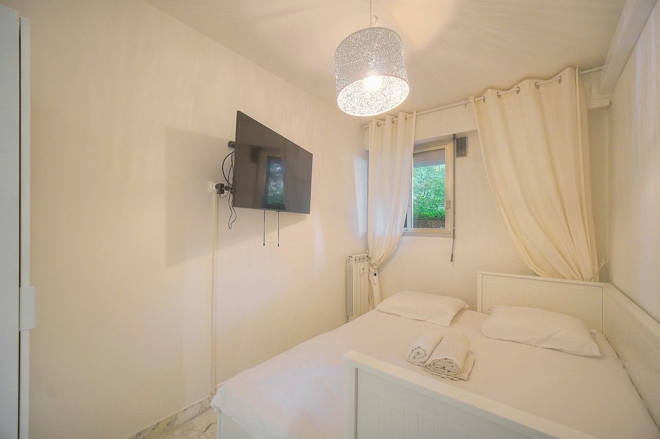 A bedroom with a bed, a TV mounted on the wall, a window with curtains, and a radiator.