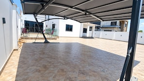 A covered patio area with tiled flooring, outdoor seating, and a view of a modern building. - ThePeppy Apartment (Prampram)