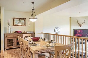 A dining area with a wooden table set for four, a wooden cabinet with glass doors, a hanging light fixture, a wall clock, and a mounted deer head. - Bear Hollow Beauty. Hottub Walk to Free Bus 5435 (Park City)