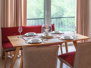 Ein Essbereich mit einem Holztisch, gedeckt für vier Personen, roten Polsterstühlen und Blick ins Grüne durch große Fenster.